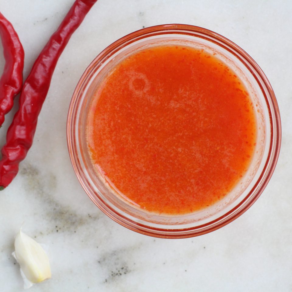 small bowl of healthy hot sauce on a surface with two chilis and a clove of garlic