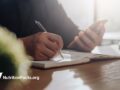 Image of man writing notes in a journal while referencing his phone screen