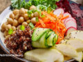 ​​Plant based dish in a bowl, containing shredded carrots, cucumber, turnips, beets, kale, apple slices, and cooked quinoa