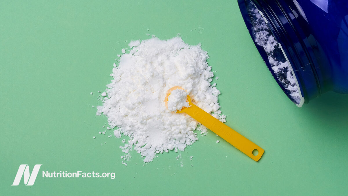 A large blue glass container on its side with spilled white taurine powder with ayellow measuring scoop in the center of the powder pile in front of a mint greenbackground
