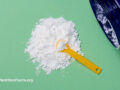 A large blue glass container on its side with spilled white taurine powder with ayellow measuring scoop in the center of the powder pile in front of a mint greenbackground