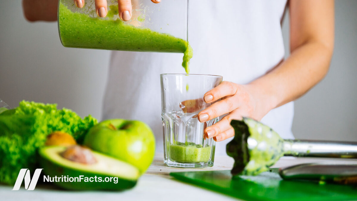 Person surrounded by vegetables and fruit, pouring a green smoothie into a glass