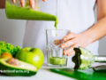 Person surrounded by vegetables and fruit, pouring a green smoothie into a glass