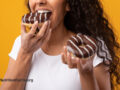 Person eating chocolate glazed doughnuts in front of a mustard yellow background