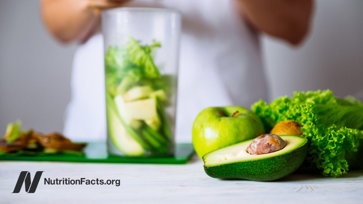 Person making a smoothie in the background with vegetables like kale and avocados